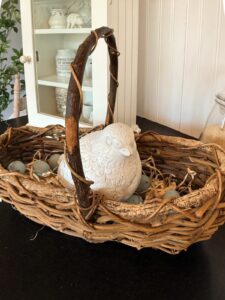 A rustic wicker basket with a curved handle holds a white ceramic bird and bluish-grey decorative eggs, creating charming vintage Easter table decor on a dark surface with a white cabinet in the background.