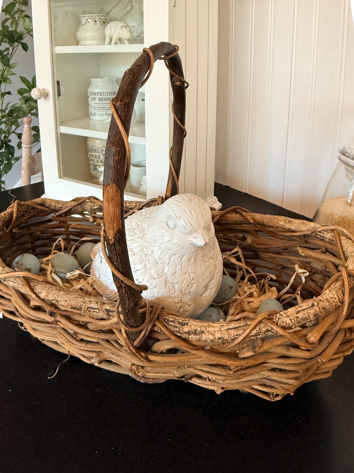 A rustic wicker basket with a curved handle holds a white ceramic bird and bluish-grey decorative eggs, creating charming vintage Easter table decor on a dark surface with a white cabinet in the background.