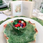 A green leaf-shaped plate holds a matching green cup filled with colorful jelly beans. Behind it is a small open storybook with an illustration, glassware, and vintage Easter table decor creating a charming leafy tablescape.