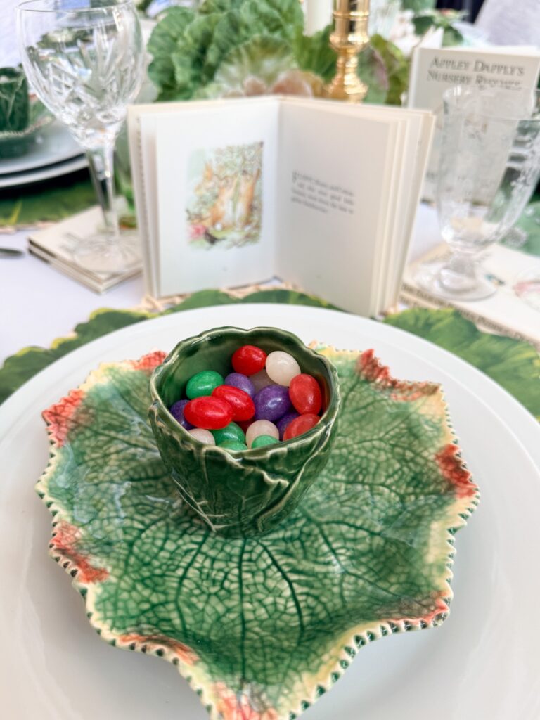 A green leaf-shaped plate holds a matching green cup filled with colorful jelly beans. Behind it is a small open storybook with an illustration, glassware, and vintage Easter table decor creating a charming leafy tablescape.