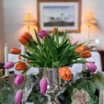 A silver vase holds vibrant orange and pink tulips on a decorated table with glassware, a cabbage-shaped plate, ceramic bunnies, and vintage Easter table decor. Two lit lamps and a framed picture hang on the white wall behind.