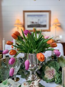 A silver vase holds vibrant orange and pink tulips on a decorated table with glassware, a cabbage-shaped plate, ceramic bunnies, and vintage Easter table decor. Two lit lamps and a framed picture hang on the white wall behind.