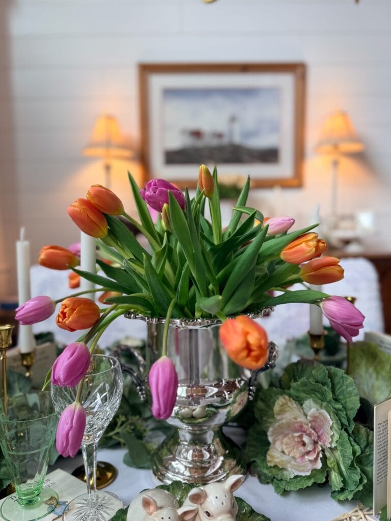 A silver vase holds vibrant orange and pink tulips on a decorated table with glassware, a cabbage-shaped plate, ceramic bunnies, and vintage Easter table decor. Two lit lamps and a framed picture hang on the white wall behind.