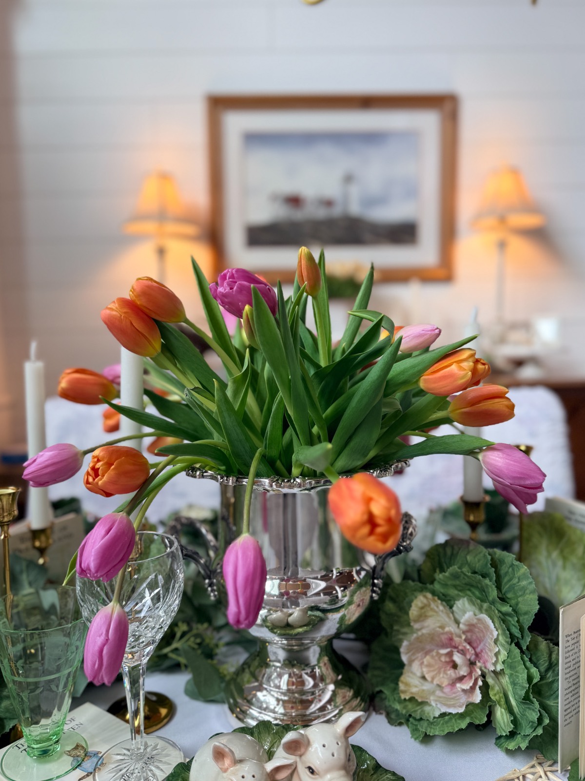 A silver vase holds vibrant orange and pink tulips on a decorated table with glassware, a cabbage-shaped plate, ceramic bunnies, and vintage Easter table decor. Two lit lamps and a framed picture hang on the white wall behind.