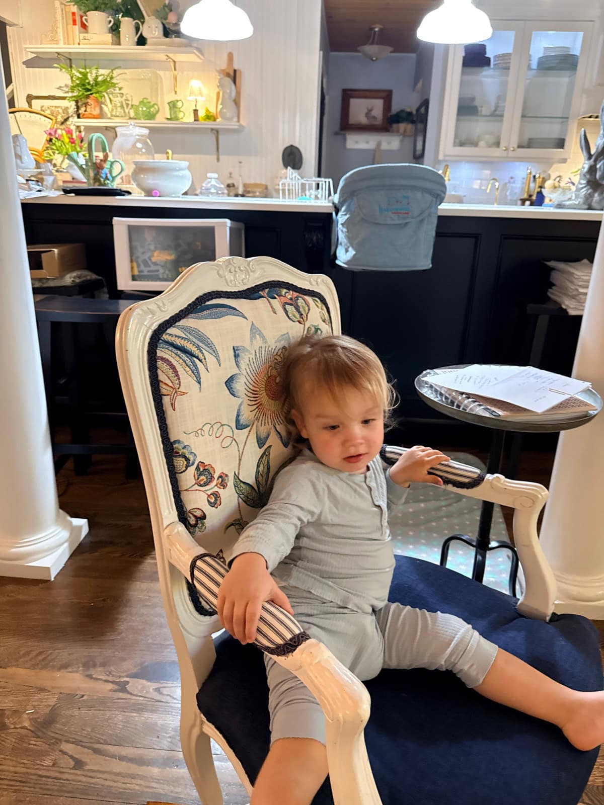 A toddler in grey pajamas sits on a decorative armchair with a floral pattern in a cozy kitchen, featuring the latest dark cabinets, white countertops, and various dishes and plants.