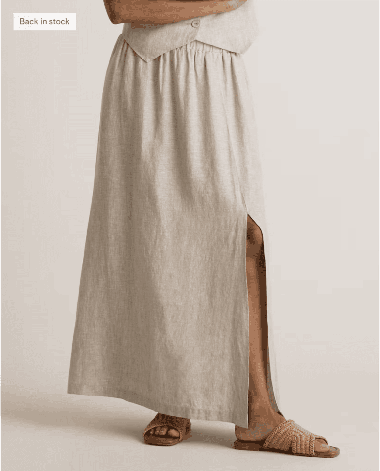 A person wearing a light beige maxi skirt with a front slit, paired with woven tan slide sandals—perfect for shopping—stands against a plain background. Only the lower half of the body is visible.