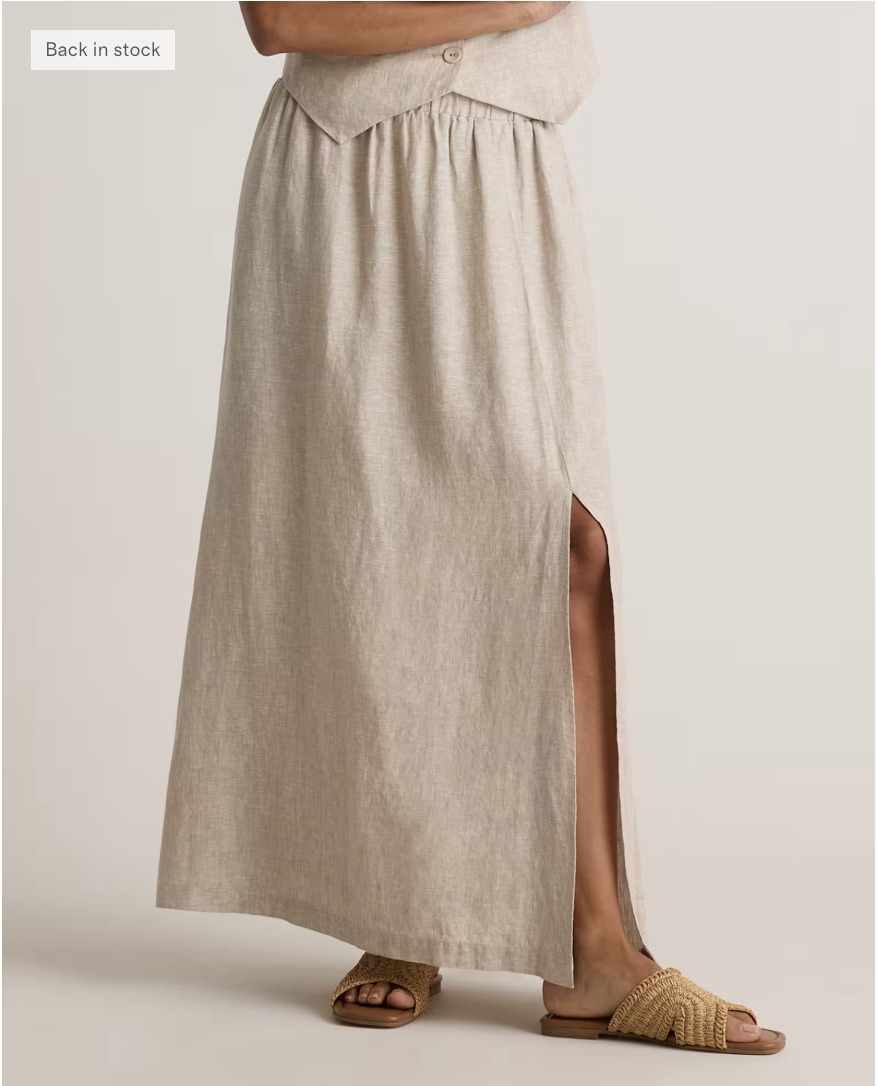 A person wearing a light beige maxi skirt with a front slit, paired with woven tan slide sandals—perfect for shopping—stands against a plain background. Only the lower half of the body is visible.