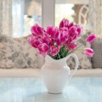 A white ceramic pitcher filled with bright pink tulips sits on a glass table in front of a sofa with the latest patterned and checkered pillows in a sunlit, cozy living room.