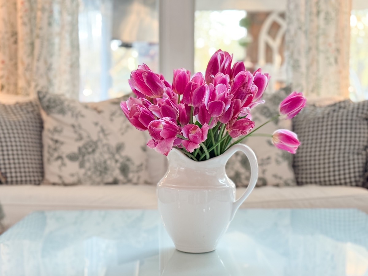 A white ceramic pitcher filled with bright pink tulips sits on a glass table in front of a sofa with the latest patterned and checkered pillows in a sunlit, cozy living room.