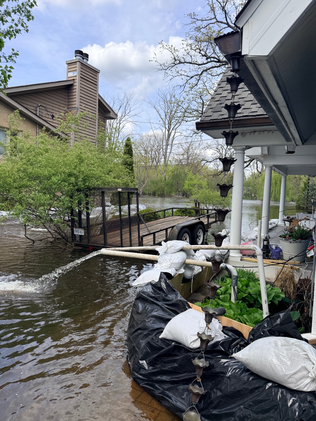Floodwaters surround a house and trailer, with the latest sandbags and plastic sheeting protecting the entrance. Plants and household items are on the porch, and a rain chain hangs from the gutter. Trees and an overcast sky appear in the background.