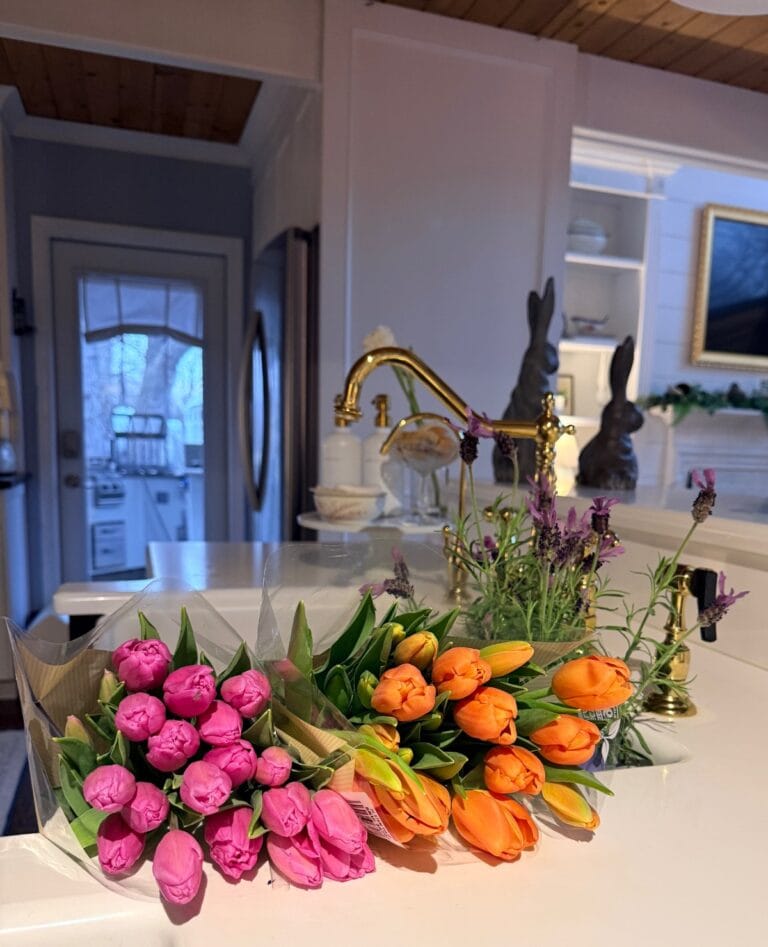 Two bouquets of tulips, one pink and one orange, rest on a white kitchen countertop—perhaps the result of shopping with friends. In the background are a gold faucet, potted lavender, bunny figurines, and a cozy, decorated kitchen.