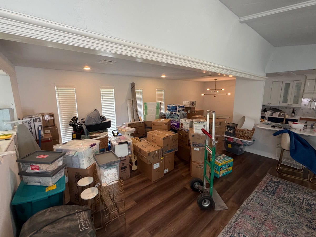 A living room filled with stacked moving boxes, packing supplies, and household items features the latest sturdy dolly nearby. The kitchen area is partially visible in the background, and the space appears ready for a move.