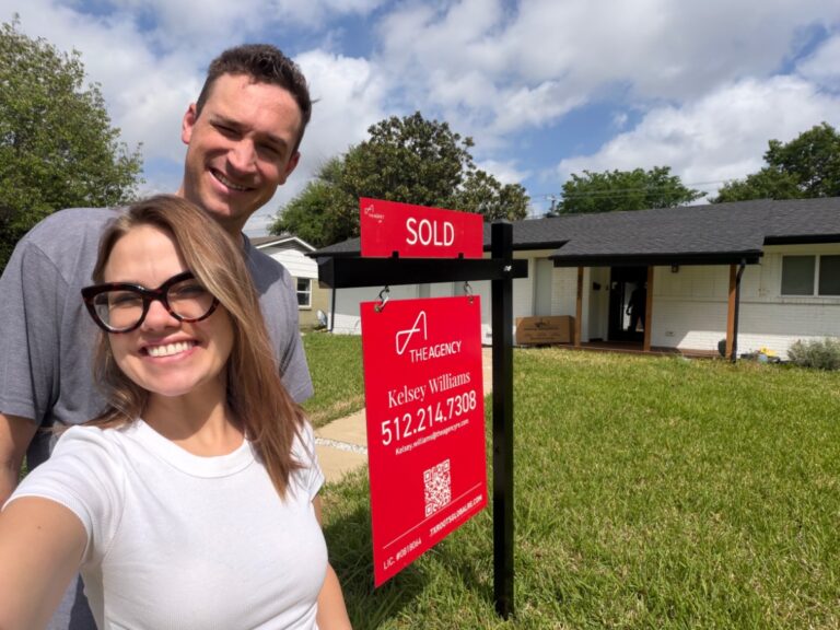A smiling couple takes a selfie in front of their latest home, with a large red "SOLD" sign in the yard. The white house features a covered porch and green lawn, marking their exciting new beginning.