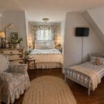 A cozy bedroom with a large bed, a small bed with a teddy bear, floral armchair, dresser with flowers, bookshelf, oval rug, wall-mounted TV featuring the latest shows, and windows with floral curtains letting in natural light.