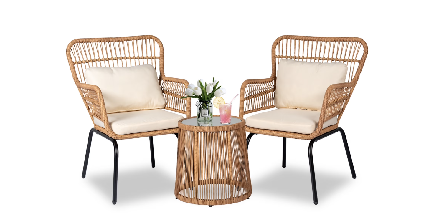 Two wicker armchairs with cream cushions face each other, separated by a round wicker side table holding a potted plant and pink drink—perfect for elevating your space or inspiring your next shopping find. All pieces are set against a white background.