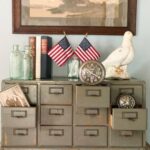A vintage metal drawer cabinet displays books, the latest glass bottles, two small American flags, an old photo, two clocks, and a white dove figurine, with a framed painting hanging on the wall above.