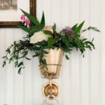 A gold wall-mounted flower pot filled with green leaves and pink, white, and purple flowers hangs above a white ceramic pitcher against a white paneled wall—a charming find for your next home shopping adventure. Part of a framed picture is visible in the top left corner.