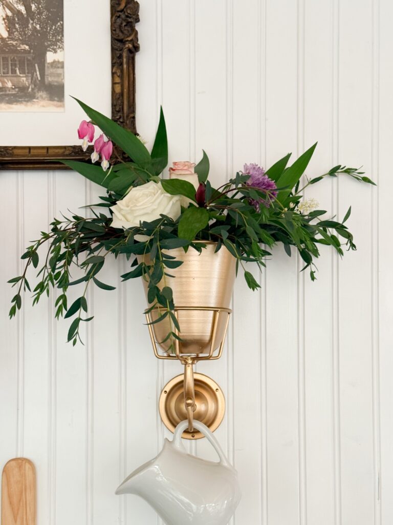 A gold wall-mounted flower pot filled with green leaves and pink, white, and purple flowers hangs above a white ceramic pitcher against a white paneled wall—a charming find for your next home shopping adventure. Part of a framed picture is visible in the top left corner.
