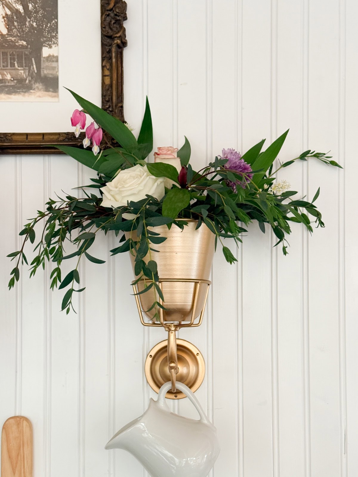 A gold wall-mounted flower pot filled with green leaves and pink, white, and purple flowers hangs above a white ceramic pitcher against a white paneled wall—a charming find for your next home shopping adventure. Part of a framed picture is visible in the top left corner.