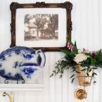 A framed vintage photo hangs above a shelf with a blue-and-white porcelain plate and pitcher, reflecting classic shopping finds. To the right, a gold wall sconce displays a small floral arrangement with roses and greenery.