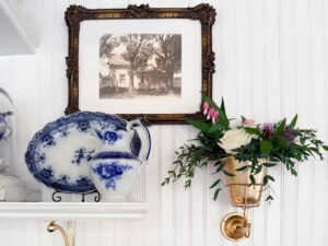 A framed vintage photo hangs above a shelf with a blue-and-white porcelain plate and pitcher, reflecting classic shopping finds. To the right, a gold wall sconce displays a small floral arrangement with roses and greenery.