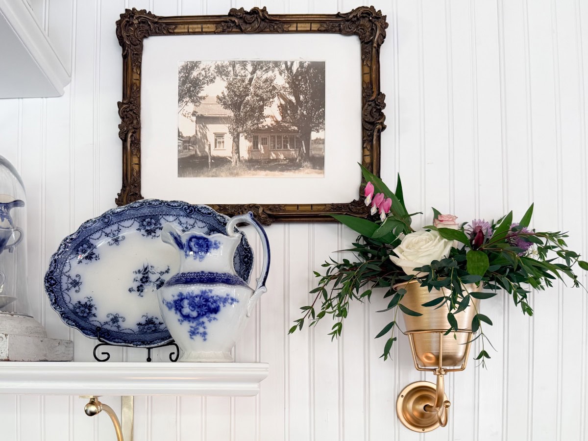 A framed vintage photo hangs above a shelf with a blue-and-white porcelain plate and pitcher, reflecting classic shopping finds. To the right, a gold wall sconce displays a small floral arrangement with roses and greenery.