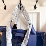 A blue CASE•MATE tote bag with wide handles wrapped in a white and blue floral-patterned scarf, perfect for shopping with friends, hangs on a black wall hook against a white paneled wall.