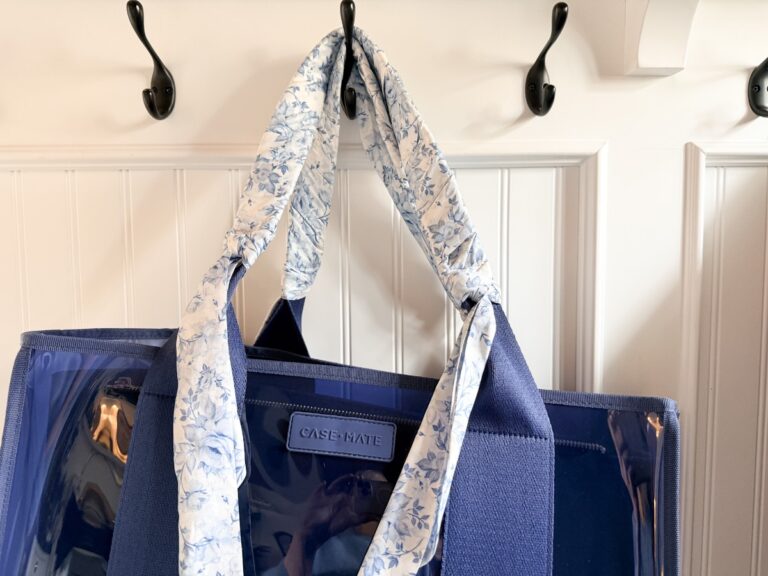 A blue CASE•MATE tote bag with wide handles wrapped in a white and blue floral-patterned scarf, perfect for shopping with friends, hangs on a black wall hook against a white paneled wall.
