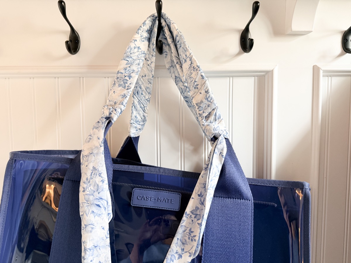 A blue CASE•MATE tote bag with wide handles wrapped in a white and blue floral-patterned scarf, perfect for shopping with friends, hangs on a black wall hook against a white paneled wall.