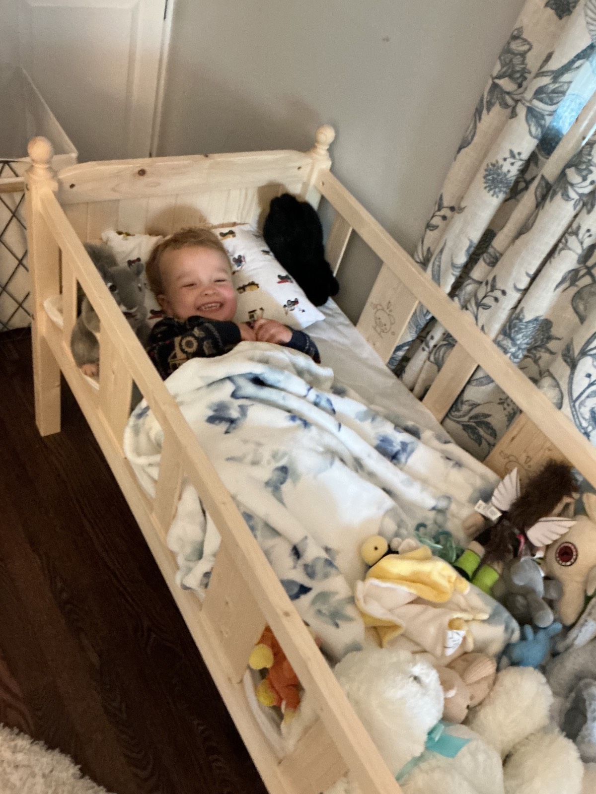 A smiling young child lies in a wooden toddler bed, covered with a white and blue blanket, surrounded by the latest stuffed animals and toys, beside a window with patterned curtains.
