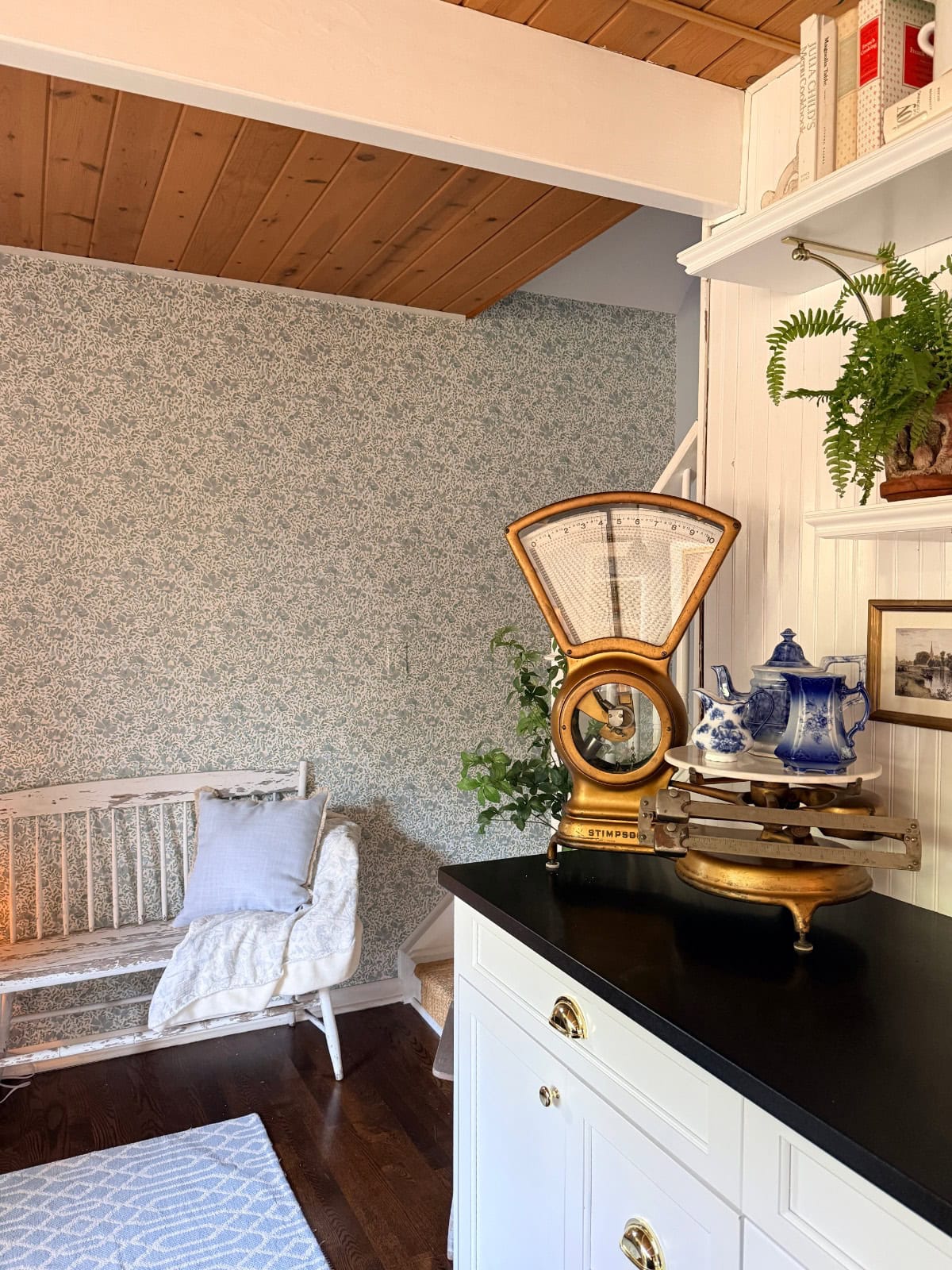 A cozy corner shows how to define an entryway with wallpaper—a vintage scale, blue and white teapot set, and potted fern rest on the counter beside a white bench, pillows, and a charming blue-patterned wall under a warm wood ceiling.