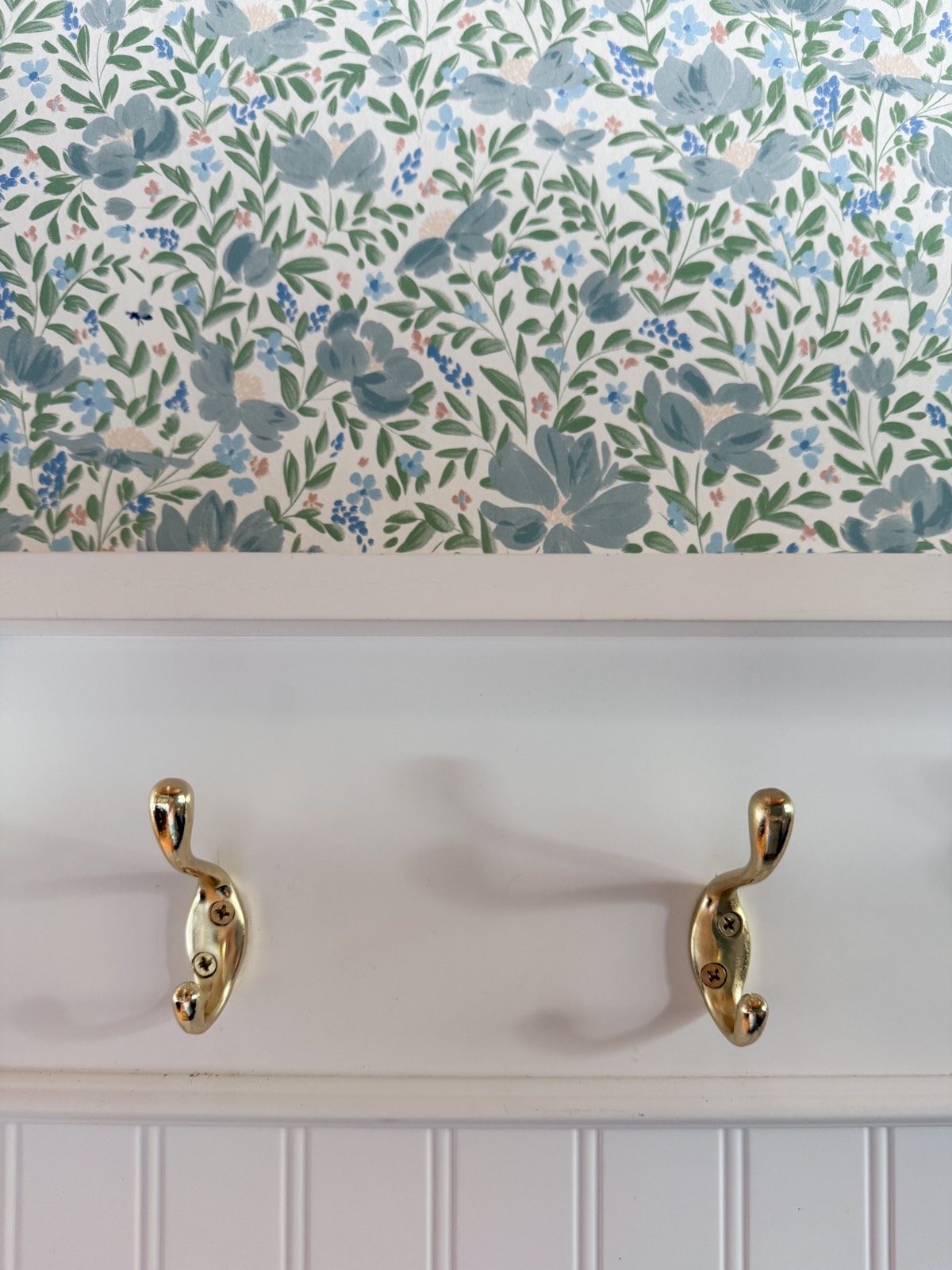 Two gold coat hooks are mounted on a white wall with blue and green floral wallpaper above, showing how to define an entryway with wallpaper. The hooks cast soft shadows over crisp white vertical paneling below.