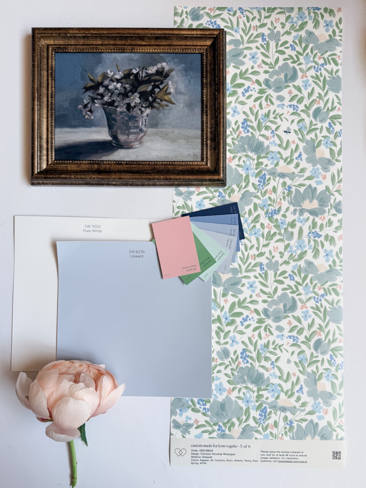 A floral wallpaper sample, paint color swatches, a white and blue paint chip, a pink peony, and a framed still life painting of flowers are arranged on a white surface—perfect inspiration for how to define an entryway with wallpaper.