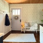 A bright entryway with floral wallpaper shows how to define an entryway with wallpaper, featuring a light blue door, white bench with a throw and pillow, patterned rug on dark wood floor, coat rack with jackets, and vintage gold scale on a white cabinet.