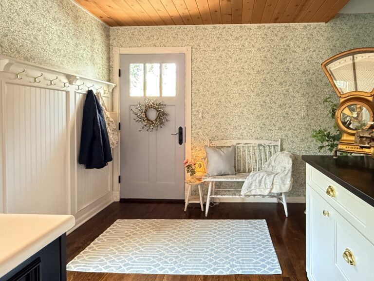 A bright entryway with floral wallpaper shows how to define an entryway with wallpaper, featuring a light blue door, white bench with a throw and pillow, patterned rug on dark wood floor, coat rack with jackets, and vintage gold scale on a white cabinet.
