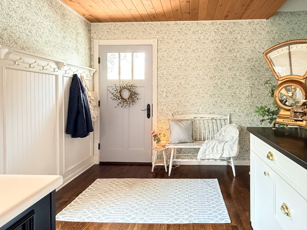 A bright, cozy entryway with a white door, small bench and throw, coat rack with jacket and bag, patterned rug, vintage scale on a white countertop—and you can learn how to define an entryway with wallpaper for added charm. Light filters in through the door window.