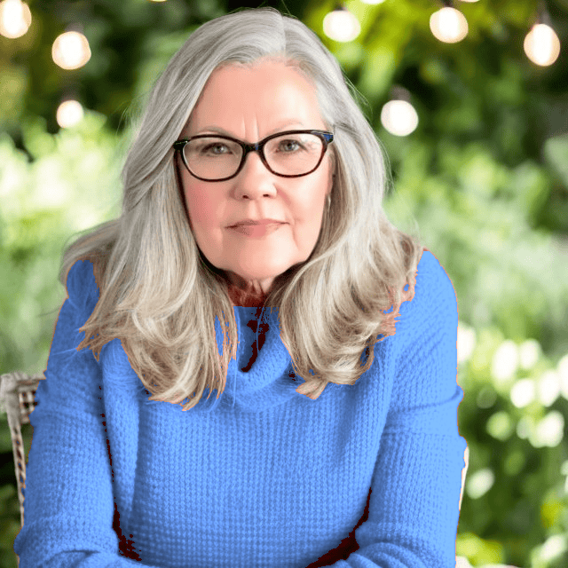 An older woman with long gray hair and glasses, wearing a textured blue sweater, sits outdoors in front of lush greenery with string lights—capturing an inviting “about me” moment.