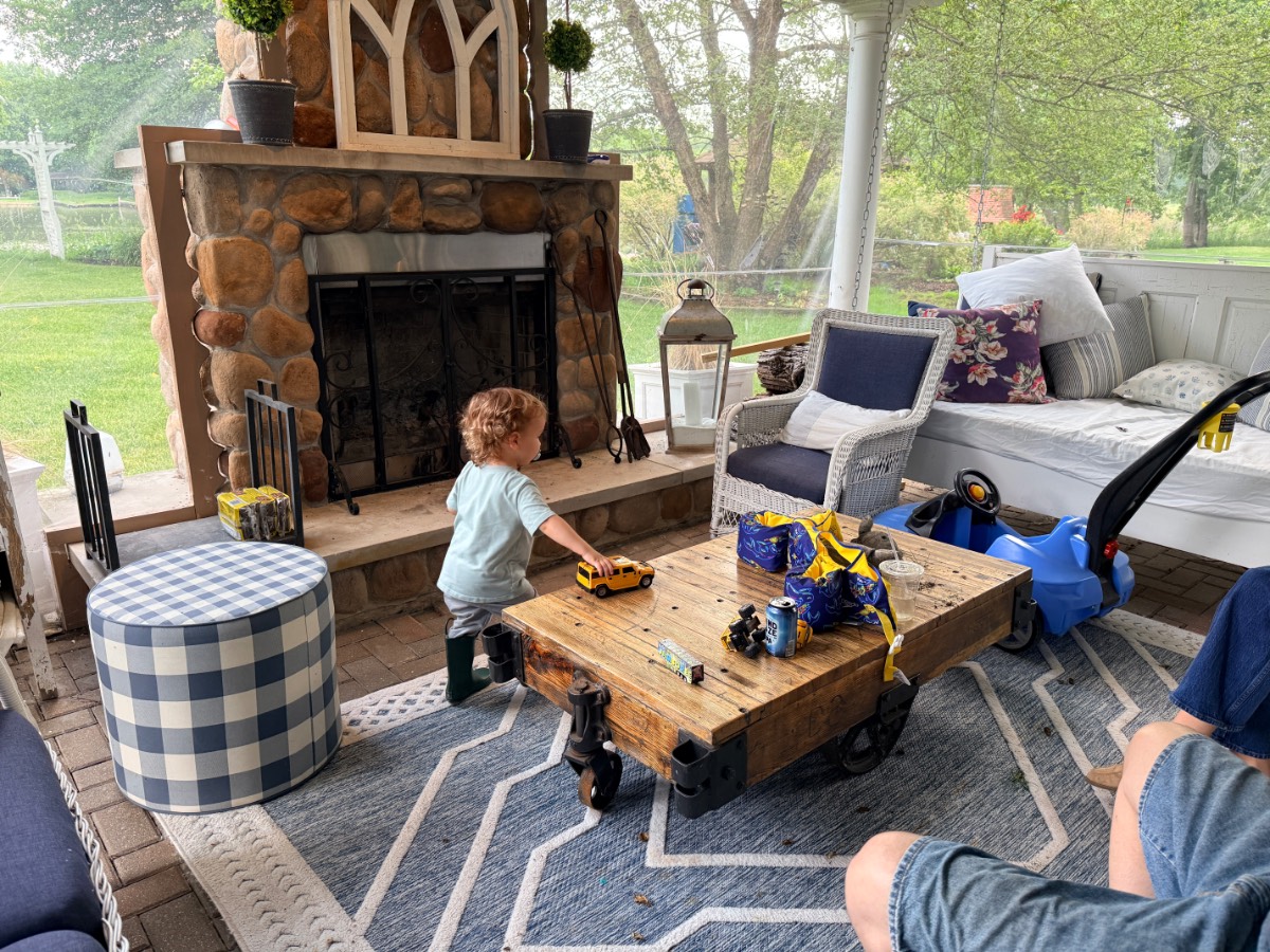 A toddler plays with the latest toy vehicles on a rustic coffee table in a cozy, covered patio. There’s a stone fireplace, cushioned seating, a checkered stool, and trees visible outside through large windows.