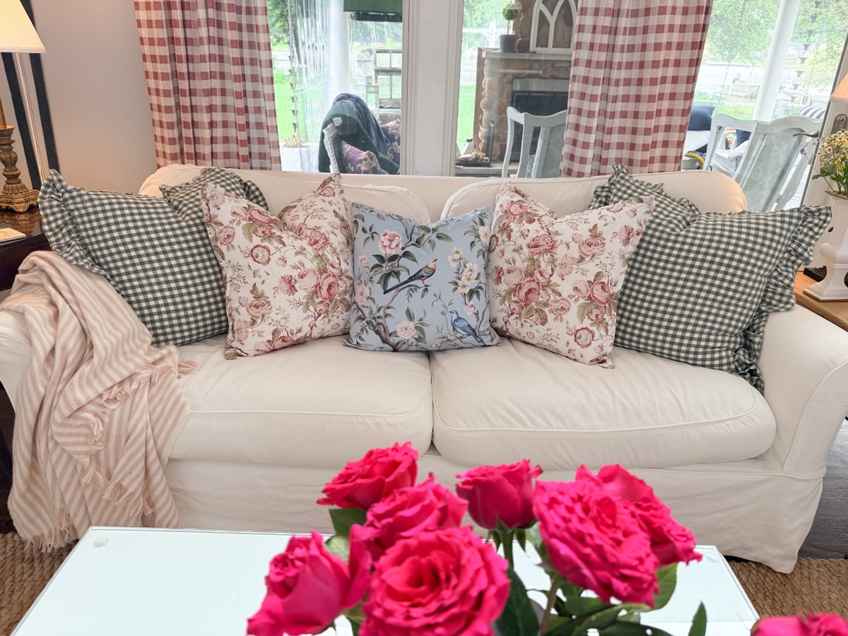 A white sofa with floral and plaid pillows shows how to refresh your home for summer with a striped blanket and pink roses.