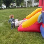 A toddler in patterned pajamas stands on grass, watching an adult slide down a yellow and red inflatable slide in a backyard near a lake, trees, and a white trellis—reminiscent of the fun energy found when shopping with friends.