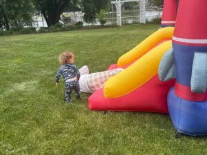 A toddler in patterned pajamas stands on grass, watching an adult slide down a yellow and red inflatable slide in a backyard near a lake, trees, and a white trellis—reminiscent of the fun energy found when shopping with friends.