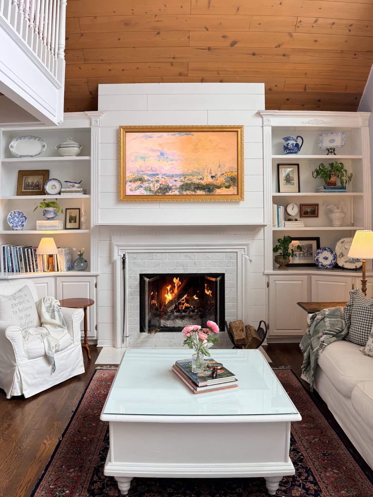 A cozy living room with a lit fireplace, white armchairs, built-in shelves with books and decor shows how to decorate a small home beautifully. A glass coffee table with flowers, wood-paneled ceiling, and warm lamps complete the inviting space.