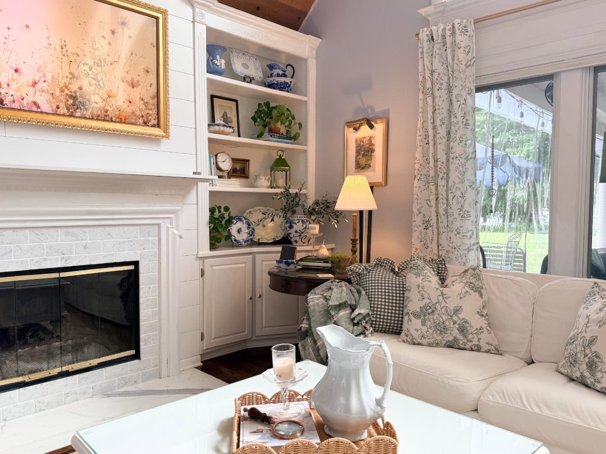 A cozy living room shows how to decorate a small home beautifully, featuring a white sofa, patterned pillows, and a glass coffee table with a silver pitcher. Built-in shelves display blue-grey decor and plants, while light streams through windows with floral curtains.