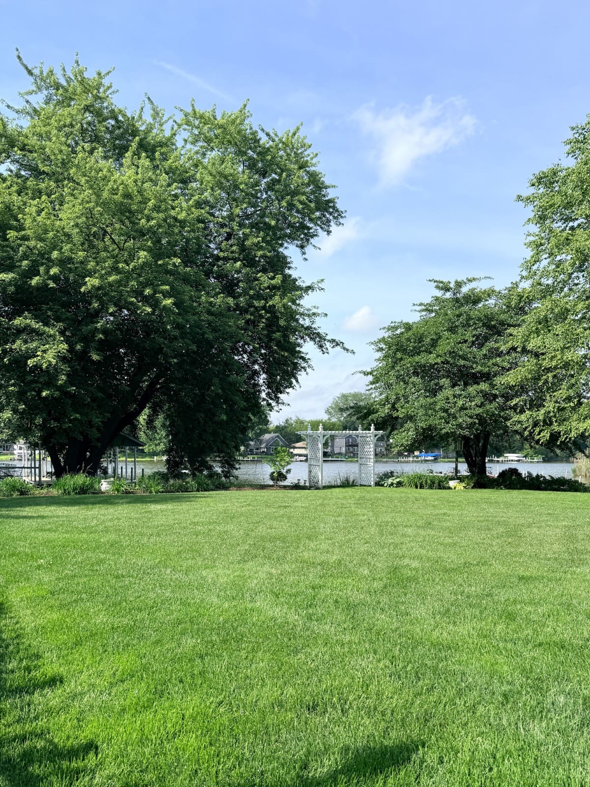 Lush green lawn with large trees and a latest white pergola near a lake under a clear blue sky.