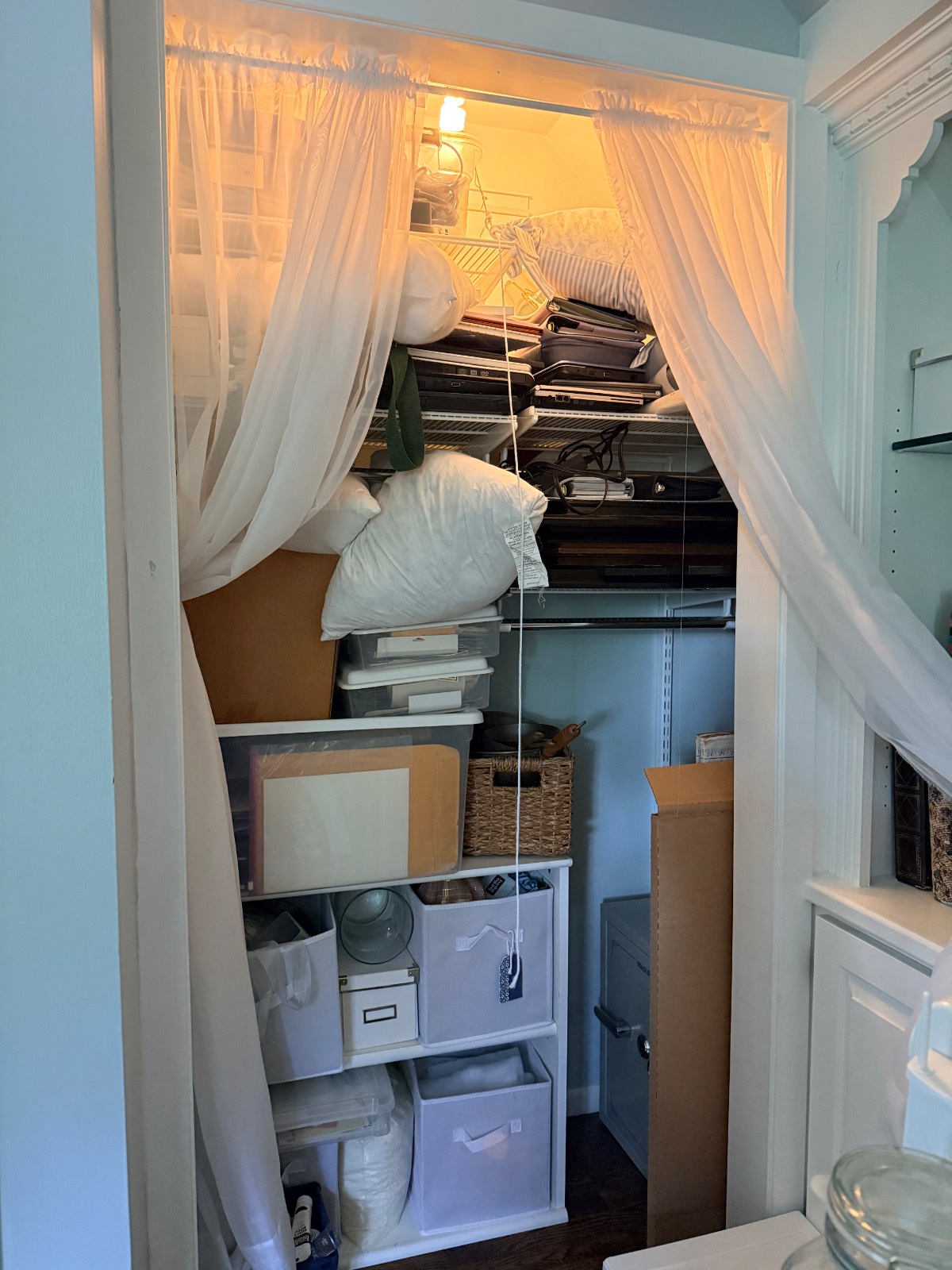 A cozy linen closet with shelves and bins, stocked with pillows, linens, baskets, and veiled in sheer white curtains.