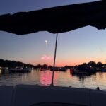 Fireworks light up the evening sky over a lake, while boats and shopping lights shimmer at sunset.