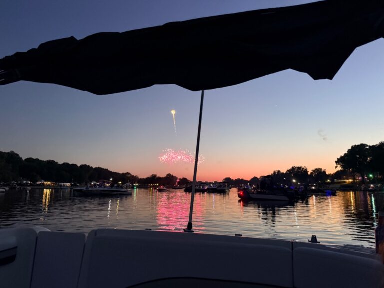 Fireworks light up the evening sky over a lake, while boats and shopping lights shimmer at sunset.