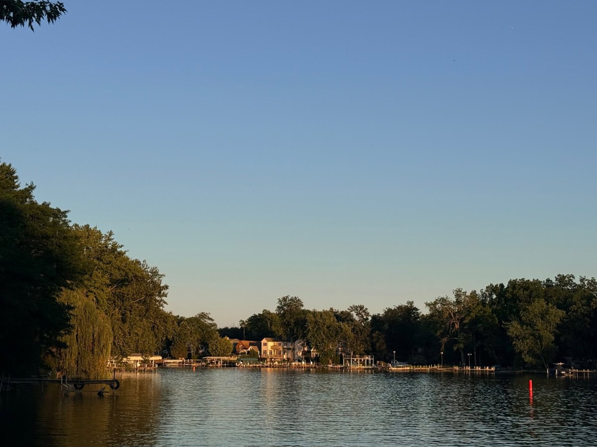 Lake view at sunset with houses, trees, docks, a red buoy, and friends shopping under a clear blue sky.