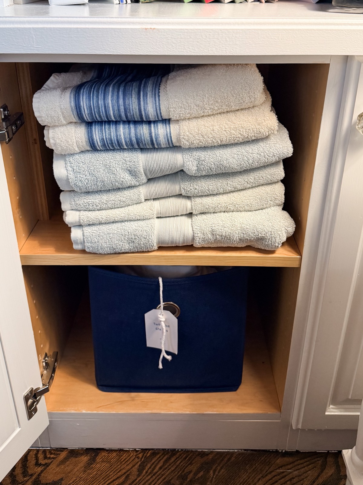 Stacked towels on a wooden shelf above a blue fabric bin in a hall linen closet with a white tag inside a white cabinet.