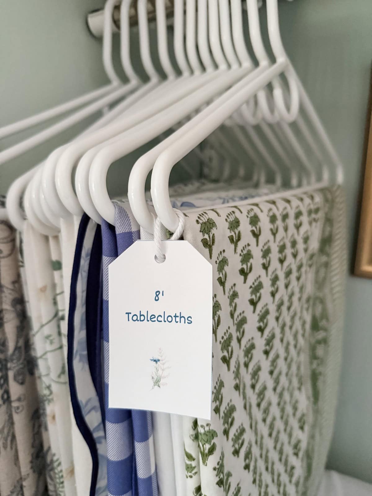 White hangers in a Hosting Closet display assorted patterned tablecloths labeled “8′ Tablecloths” with a floral tag.